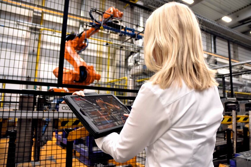 Eine Frau steht in einer industriellen Umgebung vor einem großen Robotergreifer. In ihren Händen hält sie ein Tablet, auf dem sie möglicherweise Daten überwacht oder Steuerbefehle für den Roboter eingibt. Der Fokus liegt auf der Interaktion zwischen moderner Technologie und menschlichem Eingreifen.