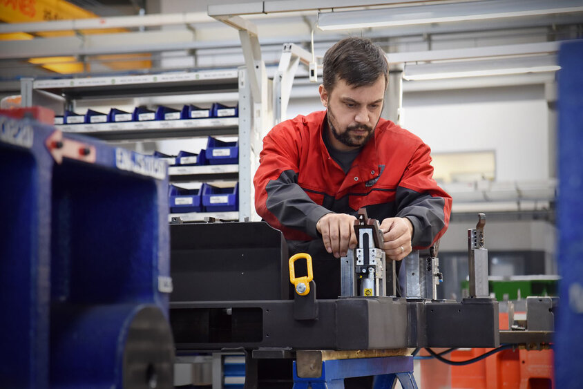 Das Bild zeigt einen Werkzeugbautechniker von weba, der mit Konzentration eine Prüfteillehre in einer produktionsnahen Umgebung zusammenbaut. Die Szene ist von Werkzeugen, Maschinen und anderen Arbeitsutensilien umgeben. Der Techniker arbeitet präzise und sorgfältig, um sicherzustellen, dass die Lehre korrekt montiert wird. Die Atmosphäre strahlt Professionalität und Effizienz aus, während der Techniker die Lehre für den Einsatz in der Produktion vorbereitet. Das Bild verdeutlicht webas übergreifende Expertise in der Umformtechnik.