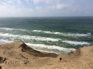 Person erklimmt eine steile Sanddüne an der dänischen Küste mit Blick auf die raue See und brechende Wellen unter bewölktem Himmel.