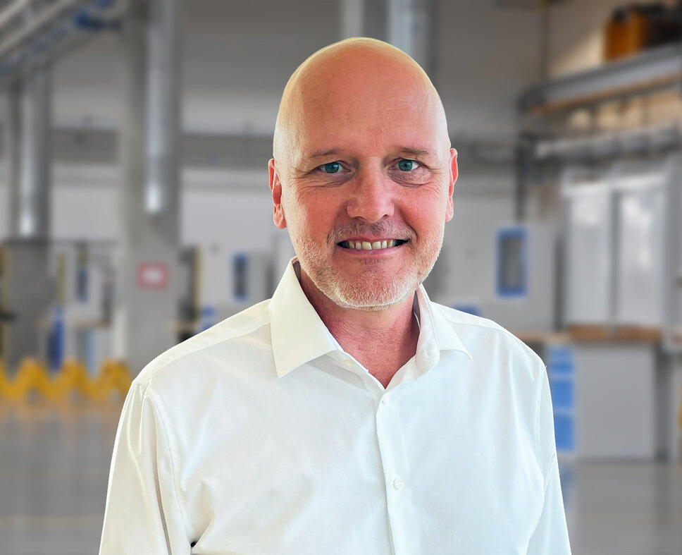Porträt von Andreas Mehlmayer in einem modernen Industrieumfeld. Er trägt ein weißes Hemd und lächelt in die Kamera. Im unscharfen Hintergrund sind helle Fertigungseinrichtungen und Maschinen erkennbar – typisch für eine präzise und saubere Produktionsumgebung.