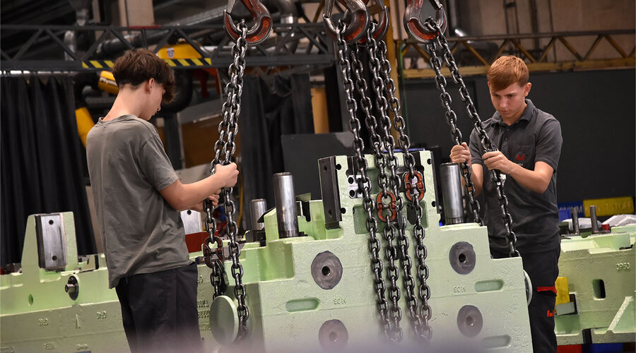 Das Bild zeigt zwei Lehrlinge in der Werkzeugbautechnik, wie sie ein Umformwerkzeug mit einem Kran spannen. Beide sind konzentriert bei der Arbeit und zeigen Teamarbeit beim sicheren Anheben des Werkzeugs. Die Szene vermittelt den Eindruck von Lernbereitschaft und praktischer Erfahrung im Umgang mit schweren Werkzeugen.