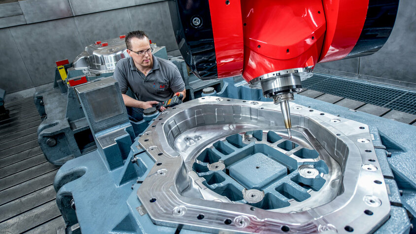 Hochpräzise Zerspanung mit modernster CNC-Frästechnologie Ein Facharbeiter bedient eine hochmoderne CNC-Fräsmaschine mit einem großen Werkzeugbauteil. Die glänzende Metalloberfläche des Bauteils zeigt präzise Zerspanungsergebnisse, während die rote Fräseinheit prominent im Vordergrund zu sehen ist.