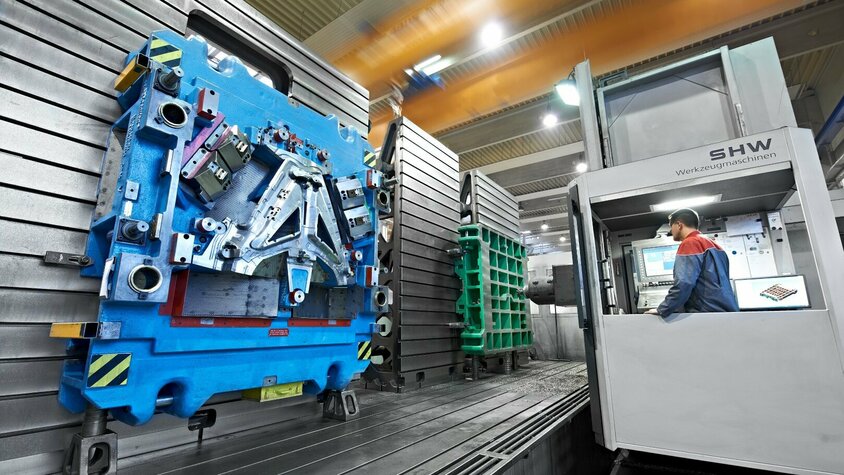 Zerspanung auf höchstem Niveau: SHW-Werkzeugmaschinen im Einsatz Ein Techniker arbeitet an einer großen SHW-Werkzeugmaschine in einer Produktionshalle. Links ist ein großes, blaues Bauteil fixiert, das für die Zerspanung vorbereitet wurde. Im Hintergrund sind weitere Bauteile und Maschinenteile zu sehen.