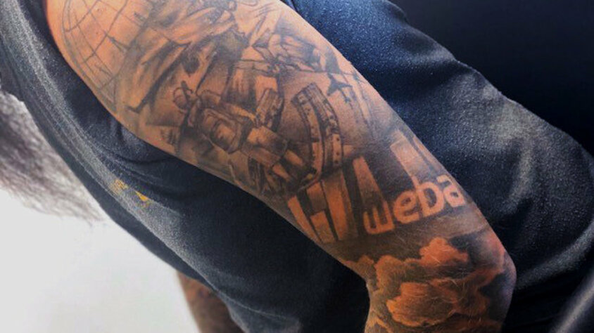 Oberarmtattoo eines weba-Mitarbeiters mit Firmenlogo und technischen Motiven Ein farblich dezentes Oberarmtattoo eines Mitarbeiters, das das Logo des Unternehmens „weba“ zeigt. Um das Logo herum sind detaillierte technische Elemente wie Zahnräder, Maschinen und eine Weltkugel abgebildet. Das Tattoo symbolisiert die enge Verbindung des Mitarbeiters zu weba Werkzeugbau und seiner Leidenschaft für Technologie und Präzision.