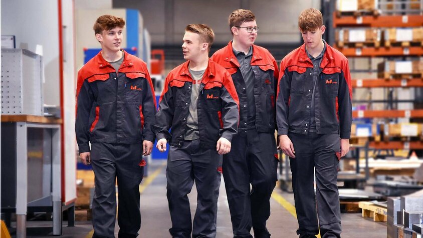 Lehre Metalltechnik - Lehrstelle in Steyr bei weba Werkzeugbau Vier engagierte Lehrlinge von Weba Werkzeugbau schlendern durch die lebhafte Produktionshalle. Sie unterhalten sich angeregt, während sie an beeindruckenden Zerspanungsmaschinen und gut sortierten Regalen mit Werkzeugen und Materialien vorbeigehen. Das Bild symbolisiert die Lehre in der Metalltechnik bei weba und vermittelt eine Atmosphäre von Lernen und praktischer Anwendung, eingebettet in die reale Arbeitsumgebung eines modernen Werkzeugbauunternehmens.