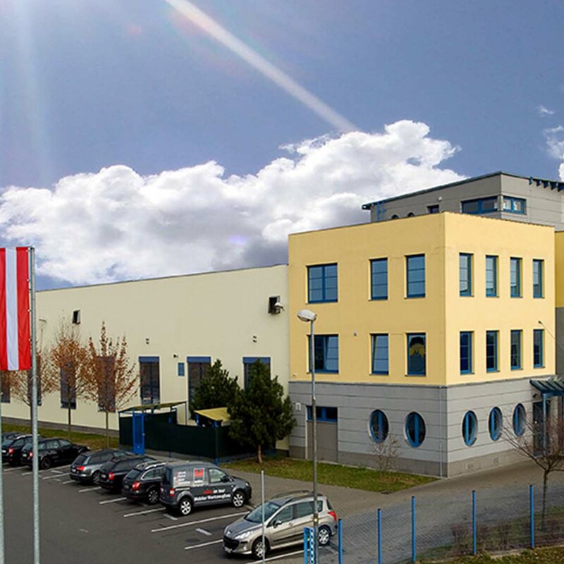 Side view of the building of the Olomouc production site from the founding days. Bright blue sky with white clouds. The building is yellow-grey in color and the flags of the European Union, the Czech Republic (CZ) and Austria (AT) fly in front of it.