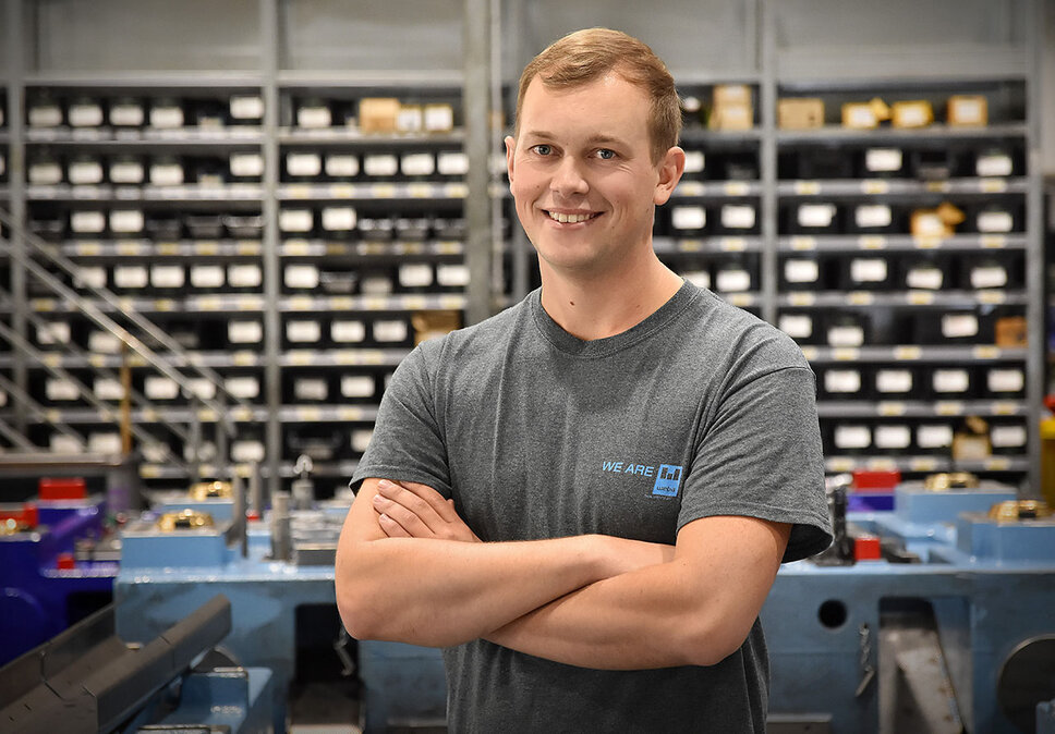Ein Werkzeugbautechniker von weba Werkzeugbau, fotografiert in der Produktionsumgebung, blickt direkt und mit einem stolzen Lächeln in die Kamera. Er steht in einer selbstbewussten Pose, die seinen Stolz auf seine Arbeit unterstreicht. Im Hintergrund sind ordentlich angeordnete Betriebsmittelregale und ein markantes blaues Umformwerkzeug zu sehen, was auf die gut organisierte und technologisch fortschrittliche Arbeitsumgebung hinweist. Das Bild vermittelt ein Gefühl von Professionalität und Zufriedenheit im Arbeitsumfeld, und hebt die persönliche Verbindung des Technikers zu seiner Arbeit und seinem Arbeitsplatz hervor. Das Bild steht symbolisch für Praktikum-Jobs in Steyr.