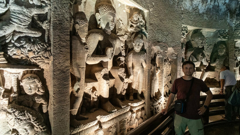 Fein gearbeitete Reliefs in einer antiken Höhle Eine Innenansicht einer Höhle mit kunstvoll gemeißelten Reliefs, die Buddha und andere Figuren darstellen, beleuchtet von weichem Licht. Eine Person steht im Vordergrund und betrachtet die beeindruckenden Schnitzereien.