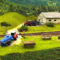 Close-up of a model railway featuring a red locomotive, a tractor, and a trailer in a detailed miniature landscape.