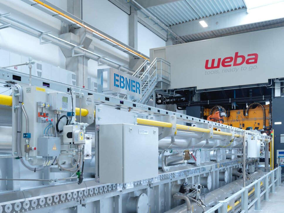 Foto einer Warmumformanlage von weba Werkzeugbau, auf dem ein 22 Meter langer Rollenherdofen und eine 1.600 Tonnen Umformpresse zu sehen sind. Die Anlage zeigt die industrielle Fertigungstechnik in Aktion, wobei der Ofen für die Erwärmung von Metallplatten vor der Umformung verwendet wird und die Presse das erwärmte Metall in die gewünschte Form bringt.