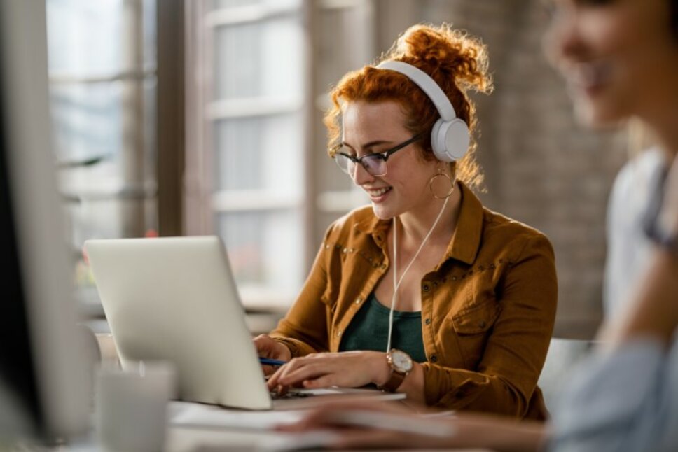 Lächelnde kreative junge Frau hört Musik mit Kopfhörern, während sie im Büro an einem Computer arbeitet.