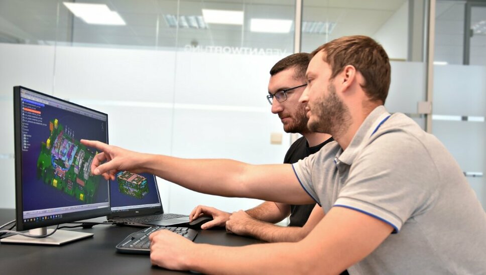 Zwei Personen sitzen vor zwei Computerbildschirmen, auf denen eine CATIA-Datei mit einer Werkzeugkonstruktion geöffnet ist. Eine der Personen zeigt auf ein spezifisches Detail des Werkzeugs auf dem Bildschirm, während sie offenbar die Konstruktion besprechen.