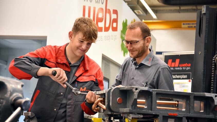 Lehre in Steyr - Ausbildung als Werkzeugbautechniker bei weba Werkzeugbau Ein Lehrling in der Werkzeugbautechnik arbeitet an einer Miniatur eines Umformwerkzeugs, während der Ausbilder aufmerksam zuschaut. weba Werkzeugbau bietet Lehre in Steyr in der Metalltechnik an.