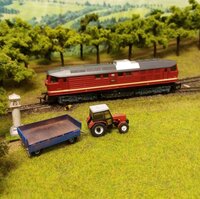 Close-up of a model railway featuring a red locomotive, a tractor, and a trailer in a detailed miniature landscape.