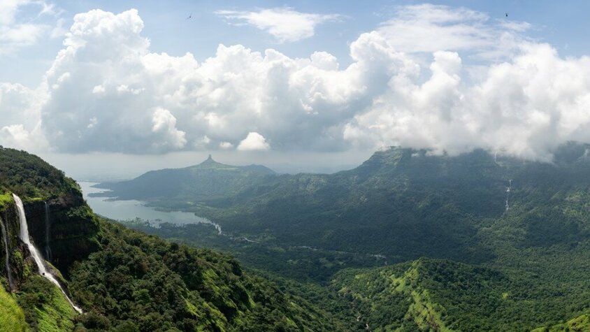 Majestätische Berglandschaft mit Wasserfällen und nebelverhangenen Hügeln Ein malerisches Panorama mit grünen Hügeln, dichten Wäldern und Wasserfällen, umgeben von einer nebligen Berglandschaft unter einem Himmel mit weißen, dramatischen Wolken.