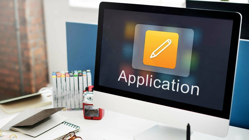 Schreibtisch mit Apple Bildschirm, Tastatur und Büromaterialien. Auf dem Bildschirm ist das Wort "Application" zu sehen, dass Symbolisch für Stellenangebote in Steyr bei weba Werkzeugbau steht. 