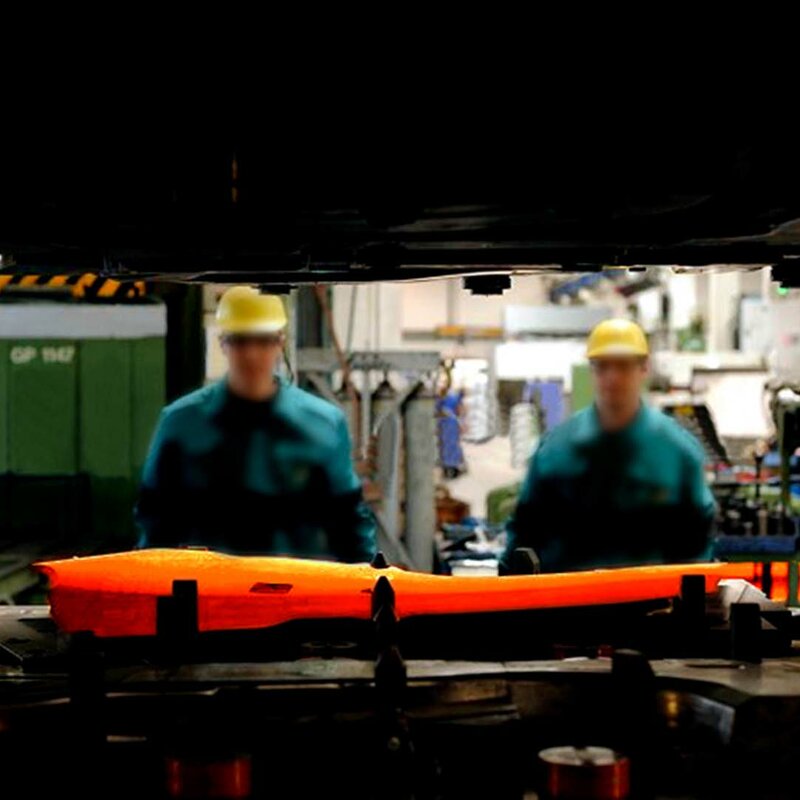 Ein durch den Spalt des Ober- und Unterteils einer Umformpresse aufgenommenes Bild fokussiert eine glühende Formplatine, die in ein Warmumformwerkzeug eingelegt ist. Im Hintergrund sind zwei Werkzeugbautechniker und die verschwommene Produktion zu sehen.