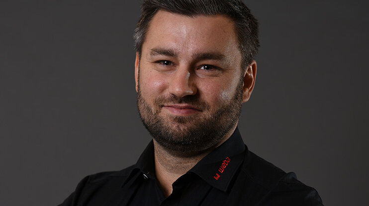 Alexander smiles confidently at the camera, wearing a black shirt with a red weba logo on the collar, posing in front of a neutral grey background.