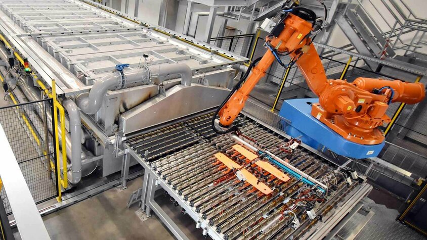 Automatisierte Warmumformung: Fortschrittliche Technologie in der Produktion Ein orangefarbener Industrieroboter entnimmt Bauteile aus einer Warmumformungsanlage. Die Anlage ist mit komplexen Fördermechanismen und Rohrleitungen ausgestattet und befindet sich in einer modernen Produktionshalle.