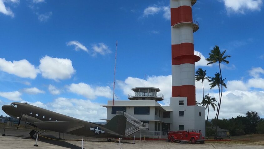 Luftfahrtkontrollturm in Pearl Harbor, Hawaii Ein markanter rot-weißer Kontrollturm in Pearl Harbor, Hawaii, mit einem historischen Militärflugzeug im Vordergrund.