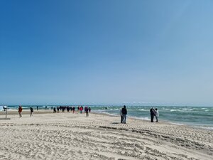 Eine Gruppe von Menschen spaziert bei sonnigem Wetter an einem Sandstrand entlang, wo sich Nord- und Ostsee treffen. Der Himmel ist strahlend blau, das Meer leicht bewegt.