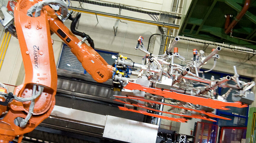 weba Werkzeugbau - Patinentransfer in der Warmumformung Ein ABB-Transferroboter ist dabei zu sehen, wie er vier glühende Formplatinen sorgfältig in ein Presswerkezug für die Warmumformung transferiert, das für die Herstellung pressgehärteter Fahrzeugbauteile verwendet wird. Die Platinen leuchten in intensivem Orange, was ihre hohe Temperatur direkt nach dem Erwärmungsprozess hervorhebt. Der Roboter, gekennzeichnet durch seine präzise Mechanik und fortschrittliche Steuerungstechnologie, bewegt sich mit einer Präzision, die für die Hochgeschwindigkeitsfertigung in der Automobilindustrie erforderlich ist. Diese Szene demonstriert die Integration von Hochleistungstechnologien in der modernen Fertigung, wo Genauigkeit und Effizienz entscheidend sind, um die Qualität und Leistung der fertigen Fahrzeugkomponenten zu gewährleisten.