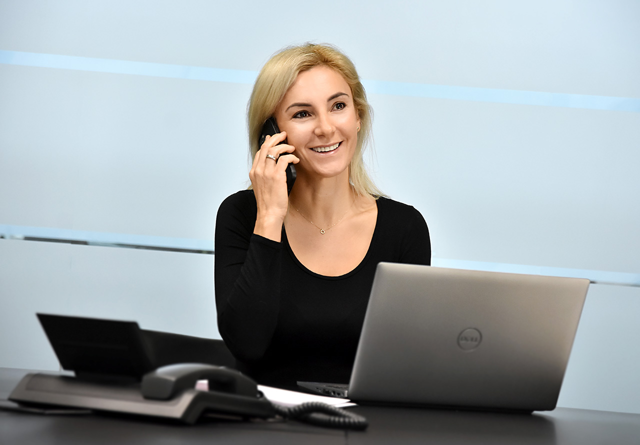 Eine weibliche Person beim Telefonieren am Handy, konzentriert und engagiert im Gespräch, mit einem aufgeklappten Laptop vor ihr auf dem Tisch, symbolisiert effizientes Multitasking und moderne Kommunikation im Arbeitsalltag.