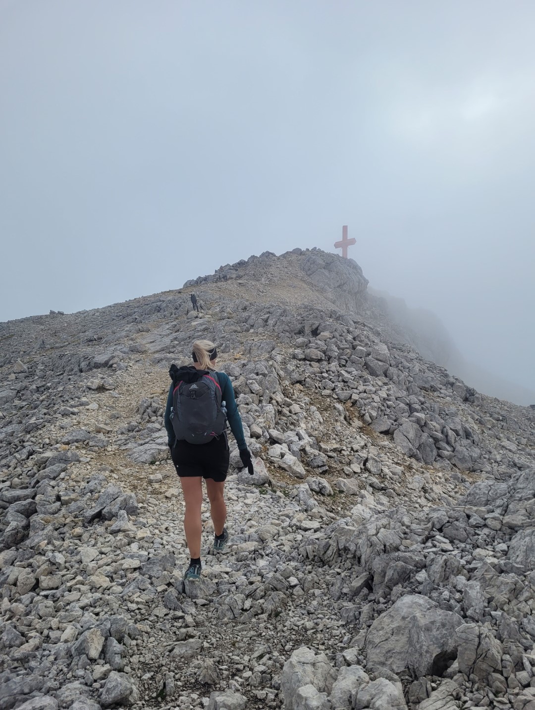 Bettina Gmainer mit Rucksack steigt einen felsigen Bergpfad hinauf, während sie sich einem Gipfelkreuz nähert. Der Himmel ist neblig, und die Atmosphäre wirkt rau und abenteuerlich.