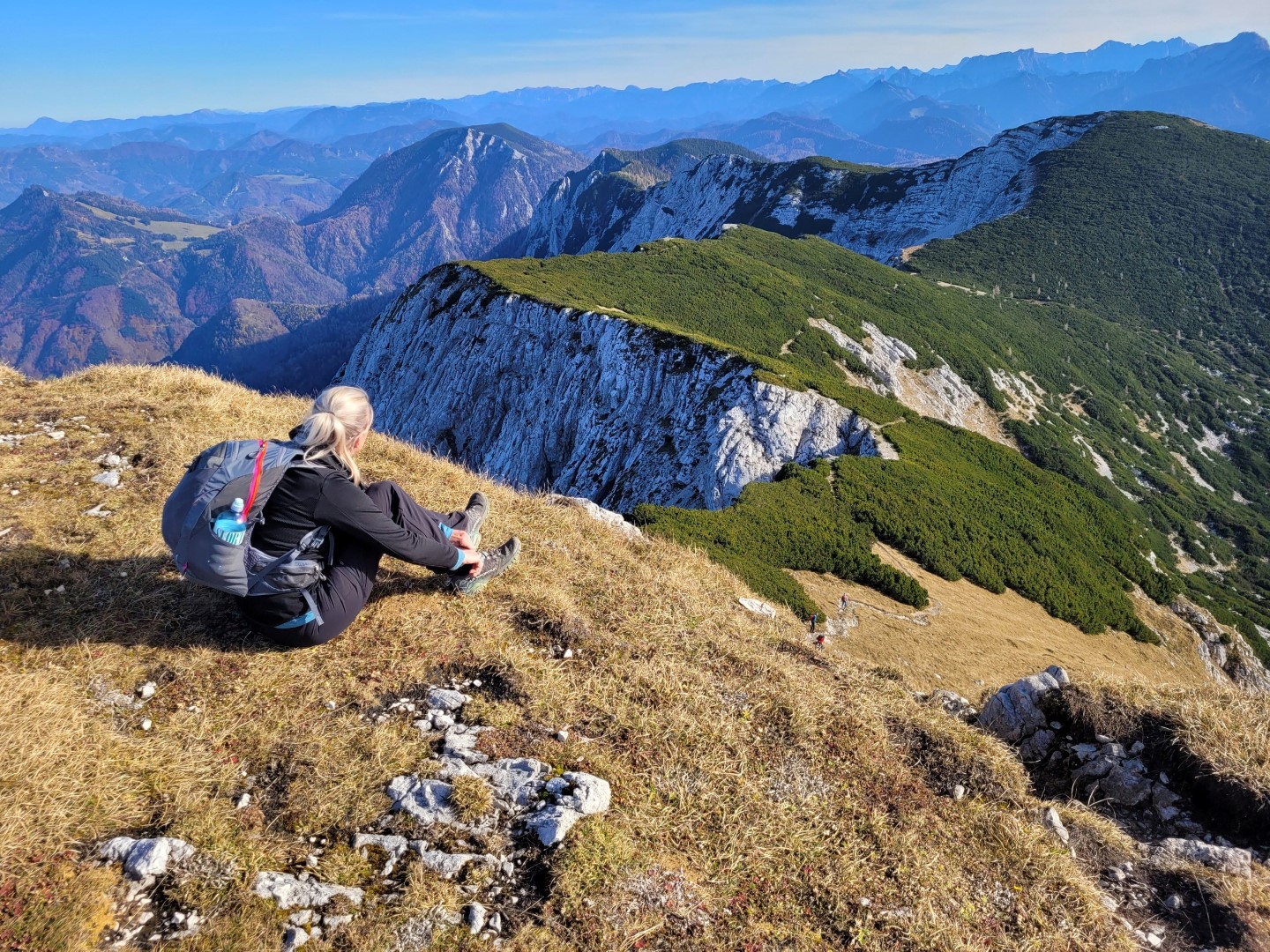 Bettina Gmainer mit Rucksack sitzt auf einer grasbewachsenen Anhöhe und blickt auf eine beeindruckende Berglandschaft mit grünen und felsigen Gipfeln.